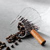 Taza medidora de café de vidrio transparente con mango de madera llena de granos de café oscuro sobre una superficie gris.