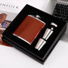 A brown leather hip flask set with stainless steel components displayed in a black box.