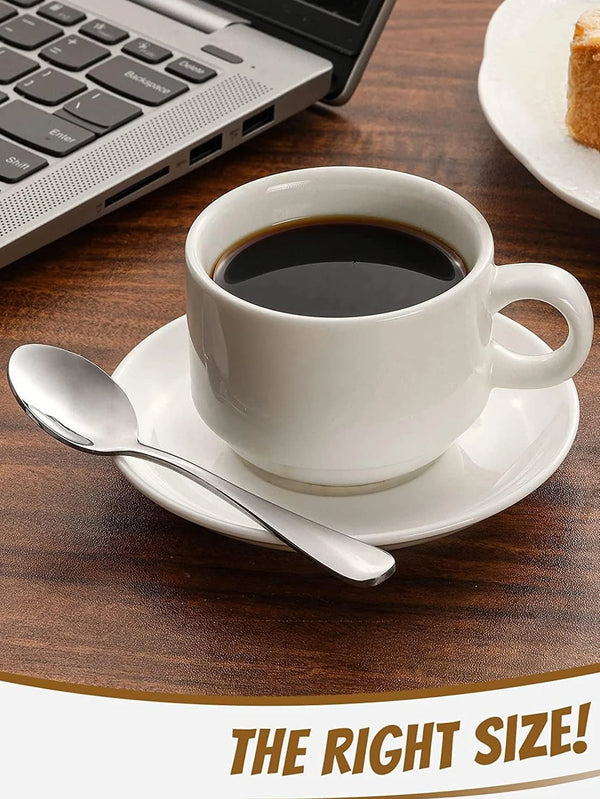 Weiße Keramiktasse und Untertasse mit schwarzer Flüssigkeit, neben einem Edelstahlrührlöffel auf Holzoberfläche.