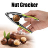 A hand is using a stainless steel nut cracker to open a walnut, with several nuts scattered on a wooden surface.