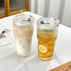 A clear drinking glass filled with an iced beverage, served with a lid and straw on a white surface.