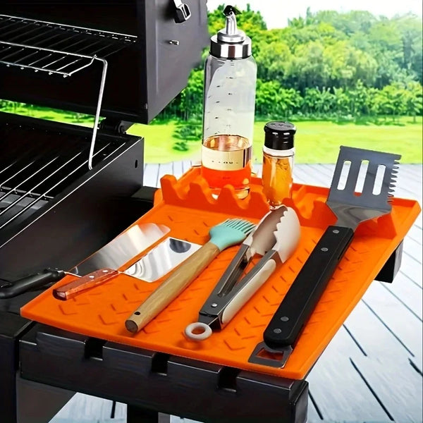 Orangener Silikonmat hält eine Glasflasche, ein Messer und verschiedene Metall- und Holzwerkzeuge auf einem schwarzen Grill.