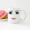 A white ceramic coffee mug featuring a stylized face with black eyes and pink lips.