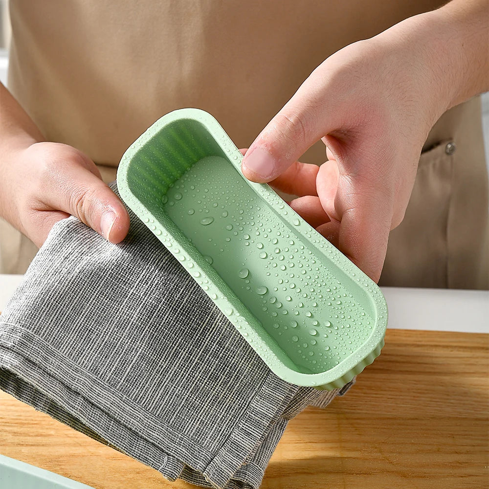 A hand is holding a silicone mini loaf baking pan in a gray cloth, showing the green silicone pan.