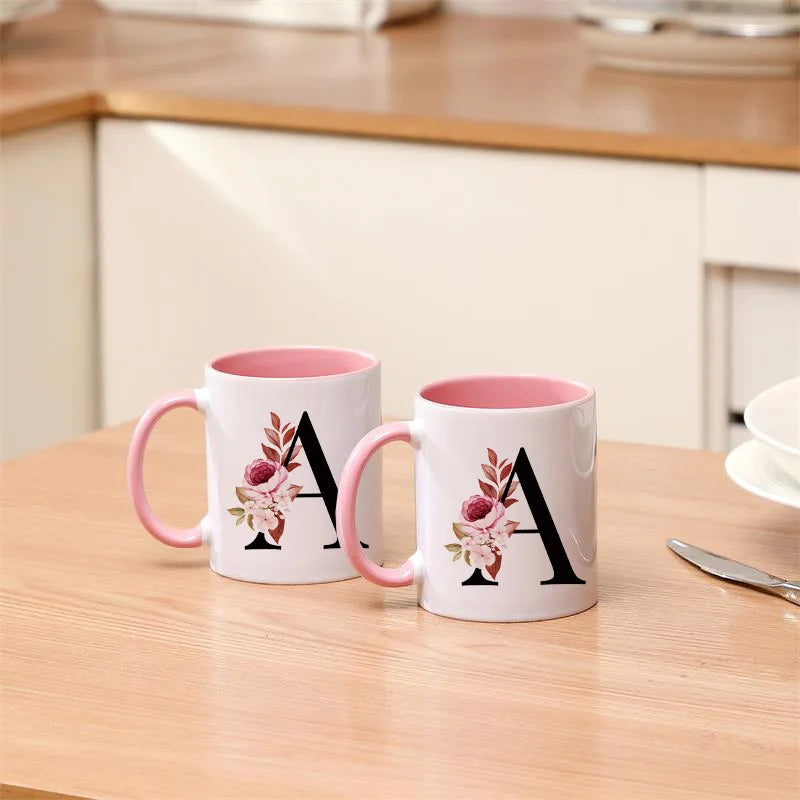 A white coffee mug with a pink handle and a large black monogram design featuring pink floral accents.