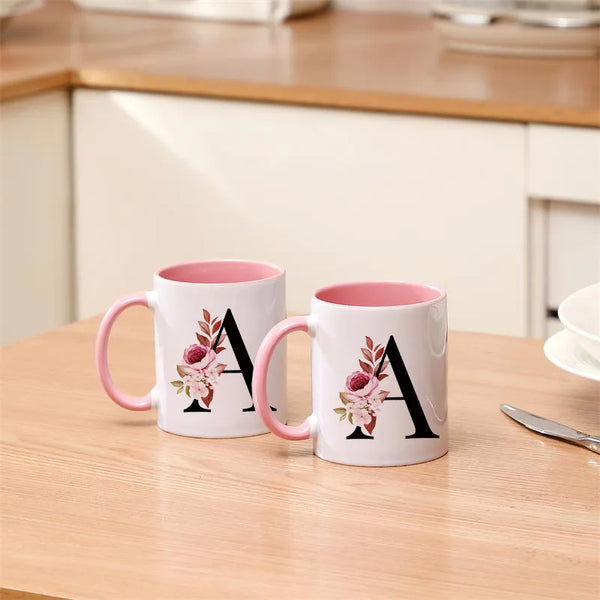 Weiße Kaffeetasse mit rosa Henkel und großem schwarzen Monogramm mit rosa Blumenakzenten.