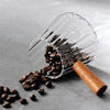 Una taza medidora de espresso de vidrio transparente con mango de madera vertiendo granos de café sobre una superficie gris.