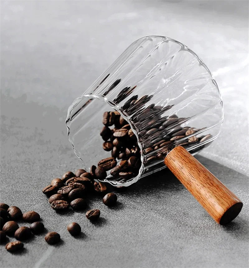 Una taza medidora de espresso de vidrio transparente con mango de madera vertiendo granos de café sobre una superficie gris.