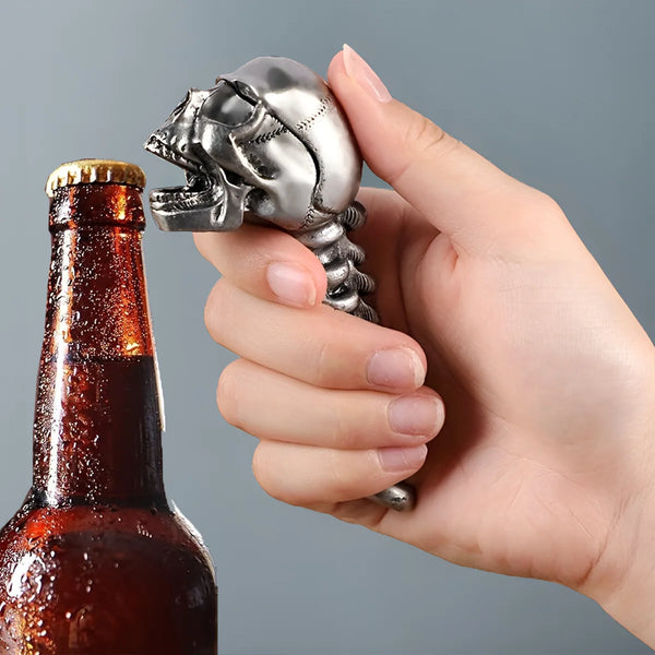 Eine Hand hält einen silbernen gotischen Schädel-Flaschenöffner neben einer braunen Glasflasche.