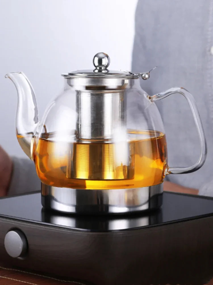 Elegant glass tea pot with a silver lid and spout pouring amber liquid on a black heating base.