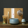 A box containing a Japanese matcha whisk set, including bamboo whisks and a metal whisk, resting on a wooden surface.