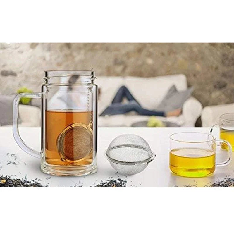 A glass mug filled with amber tea, a stainless steel mesh tea infuser, and a small glass cup on a white surface.