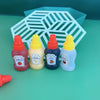 Four mini squeeze condiment bottles in orange, yellow, purple, and blue set against a light green background with a white geo