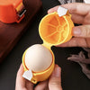 A hand holds an orange portable egg cracker and separator, opening it to reveal a whole white egg.