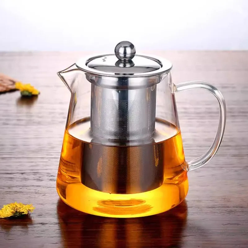 A clear glass teapot with a stainless steel infuser is filled with golden-colored tea on a wooden surface.