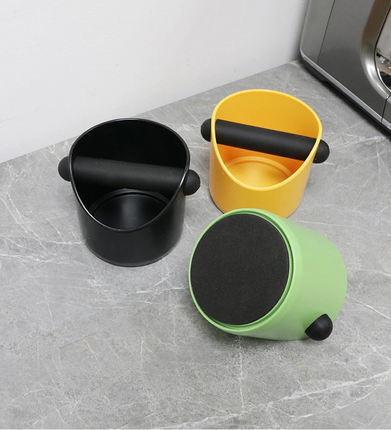 A black coffee grounds container next to a yellow one and a light green one on a gray surface.