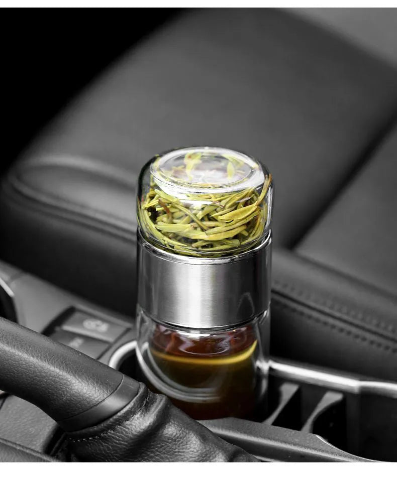 A double-layer borosilicate glass tea infuser filled with dried green tea leaves, resting on a black car interior surface.