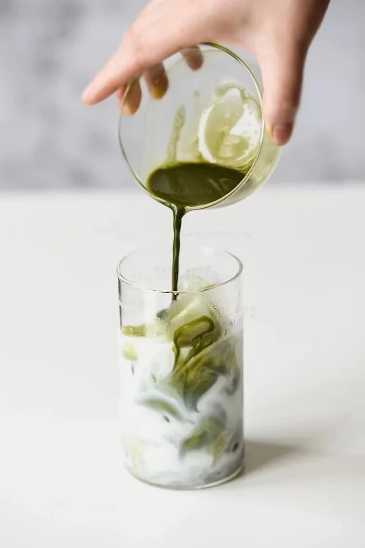 A hand pours a green liquid into a glass filled with iced milk tea.