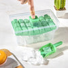 A hand is placing ice cubes into a clear plastic tray with a green lid and storage box on a white surface.