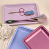 A pink tray holds a pair of scissors, a blue sponge, and a green brush on a light background.