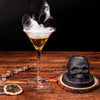 A glass of amber cocktail smoke with a skull-shaped smoker kit and citrus slice on a wooden surface.