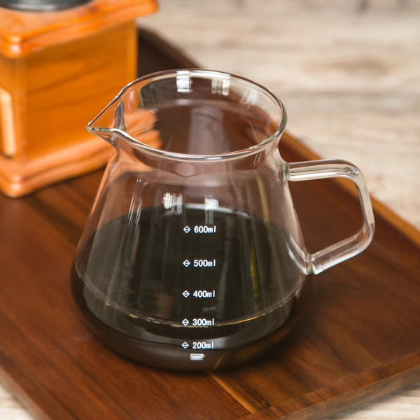 Klarer Glas-Kaffeekännchen mit Volumenmarkierungen auf Holzbrett neben einem Holz-Kaffeemühle.