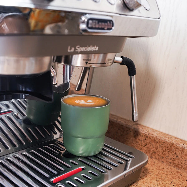 A close-up of a DeLonghi espresso machine with a glass cup containing coffee on a stainless steel tray.
