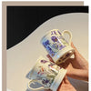 A hand holds a ceramic coffee mug with purple and green floral patterns against a light background.