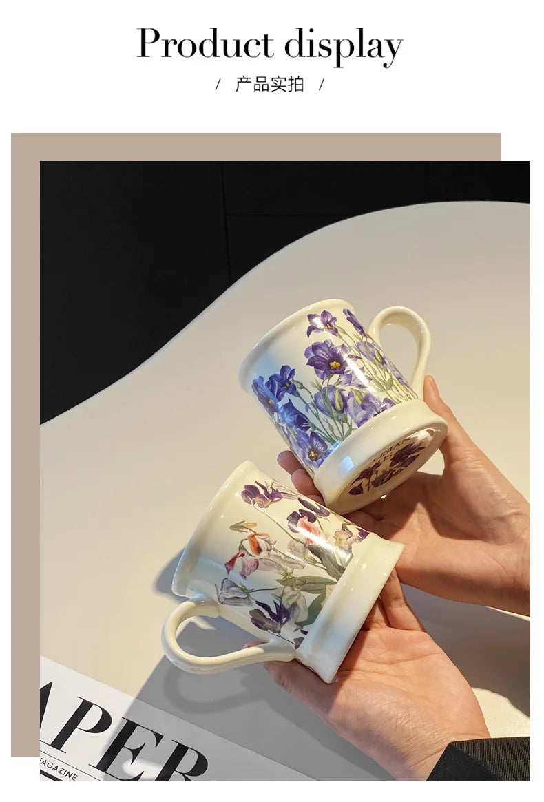 A hand holds a ceramic coffee mug with purple and green floral patterns against a light background.