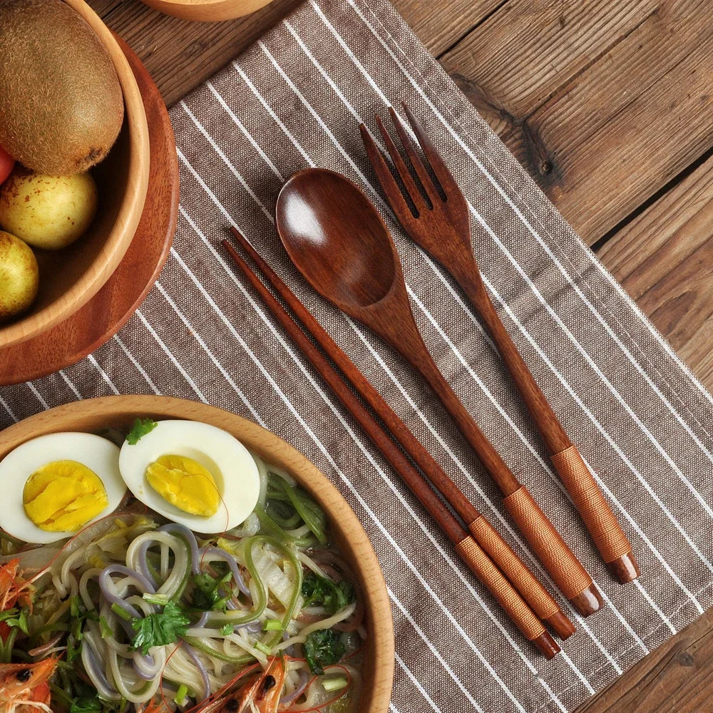 A set of natural wood dining utensils including a spoon, fork, and chopsticks resting on a striped cloth.