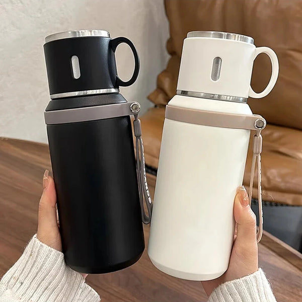 A person holds a black and silver insulated stainless steel travel mug against a brown leather sofa.