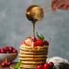 A hand pours golden syrup onto a stack of pancakes topped with fresh strawberries and cherries.