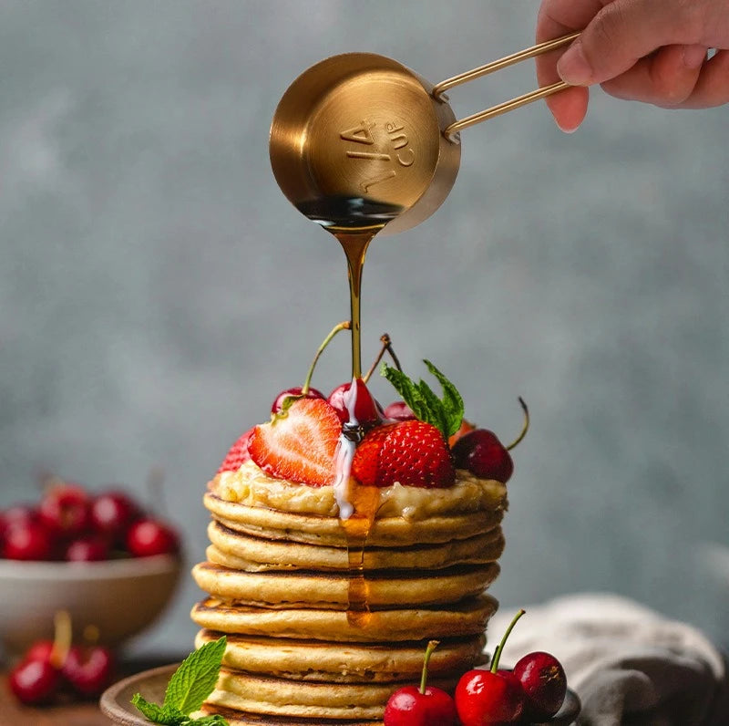 A hand pours golden syrup onto a stack of pancakes topped with fresh strawberries and cherries.