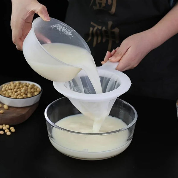 A hand pours a white liquid from a multi-mesh fine strainer into a glass bowl.