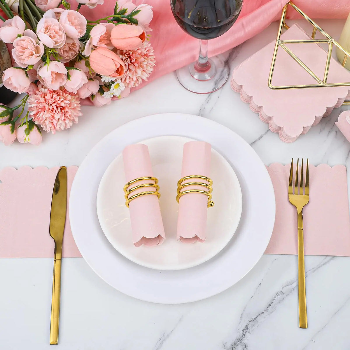 Pink rolled dessert napkins with gold string accents displayed on a white plate next to a pink floral arrangement and wine gl