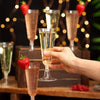A hand holds a clear disposable champagne flute filled with pale yellow liquid against a dark background with bokeh lights.