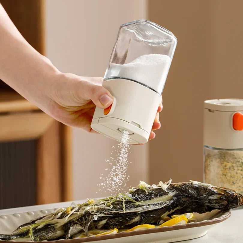 A hand is dispensing salt onto a fish on a plate using a glass dispenser bottle.