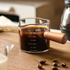 Vaso medidor de vidrio lleno de líquido oscuro etiquetado Sucafina y aprox. 75ml, sobre una superficie de madera.