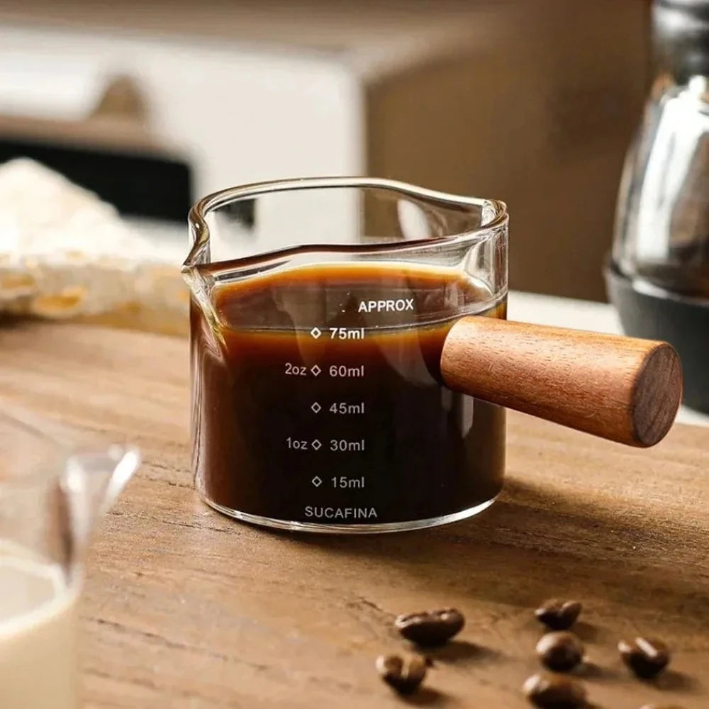 Vaso medidor de vidrio lleno de líquido oscuro etiquetado Sucafina y aprox. 75ml, sobre una superficie de madera.