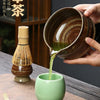 A hand pours green matcha liquid from a dark ceramic bowl into a light green cup.