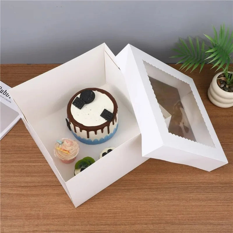 White square disposable cake boxes with a cake and greenery displayed on a wooden surface.