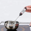 A hand holds a digital meat thermometer with a BBQ probe measuring temperature in a stainless steel bowl.