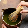 A hand is holding a bamboo whisk used for preparing matcha in a dark ceramic bowl.