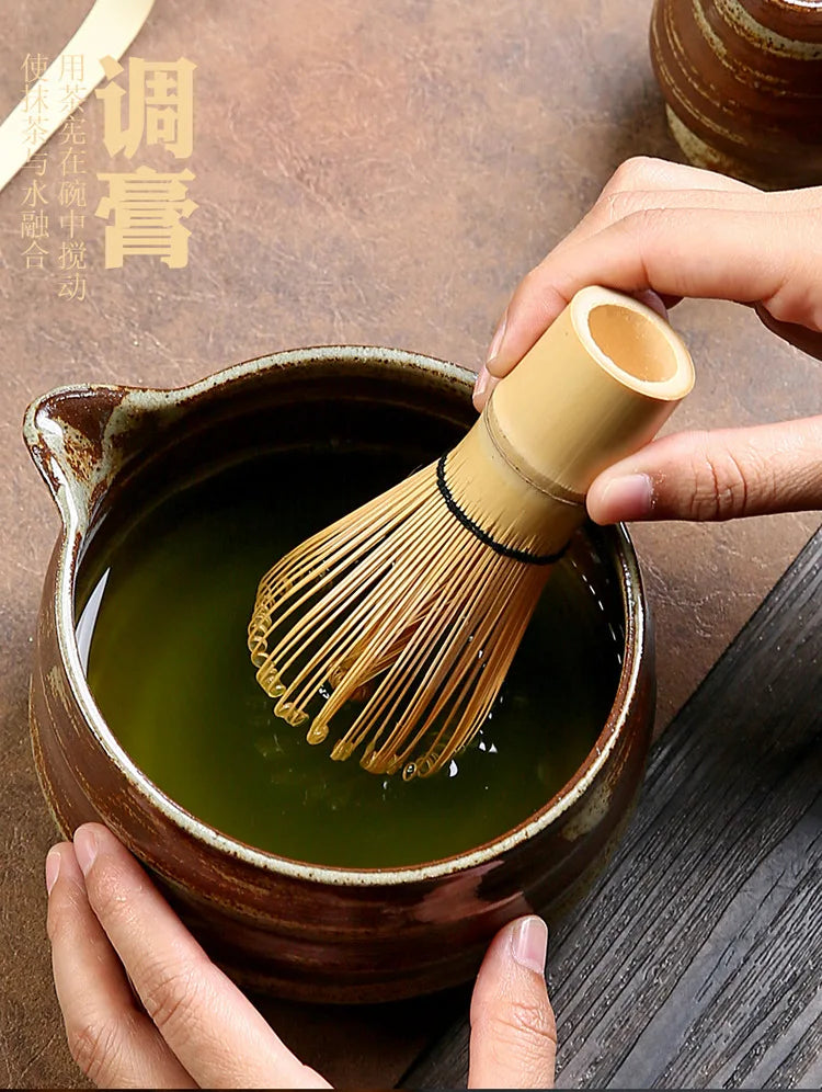A hand is holding a bamboo whisk used for preparing matcha in a dark ceramic bowl.
