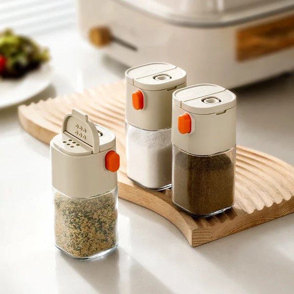 A glass dispenser bottle for salt, featuring two stacked containers with white lids and orange accents, resting on a wooden b