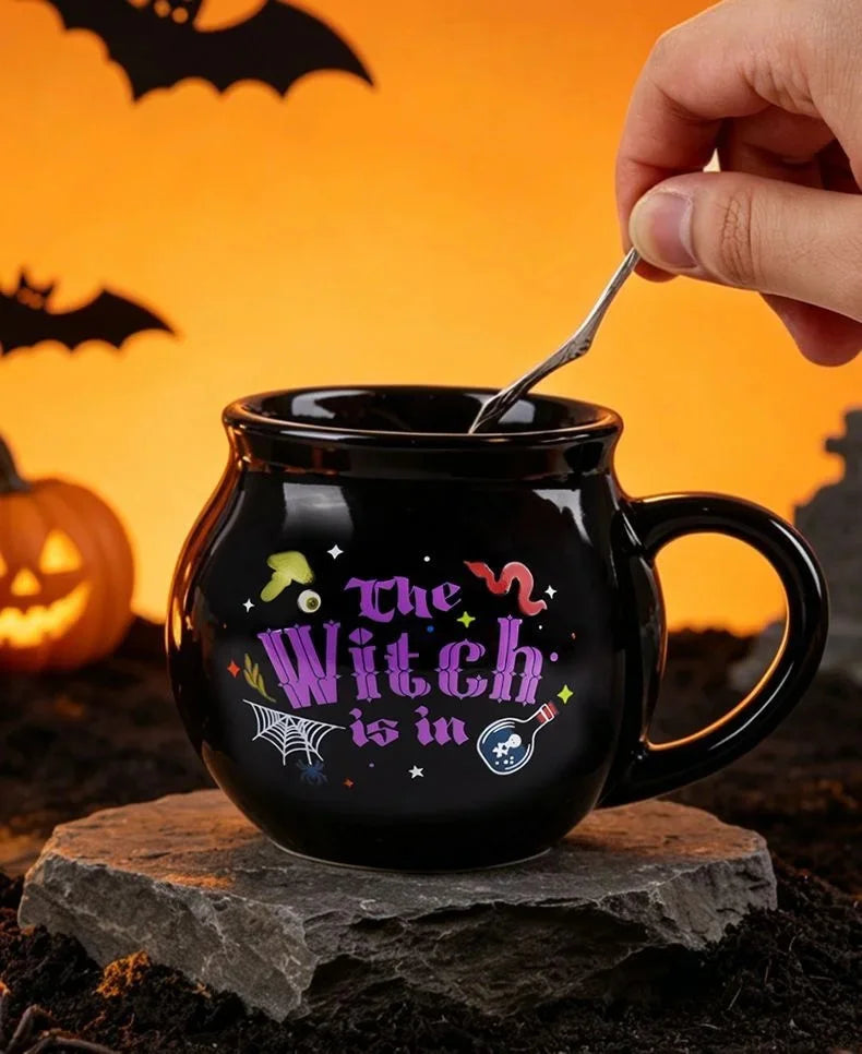 A black cauldron mug decorated with a witch theme sits on a rock, being lifted by a silver spoon against an orange background