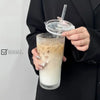 A hand holds a glass of iced coffee or milk tea with a clear lid and straw, set against a dark background.