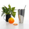 A stainless steel drinking glass filled with orange juice and a cleaning brush, next to a whole orange.