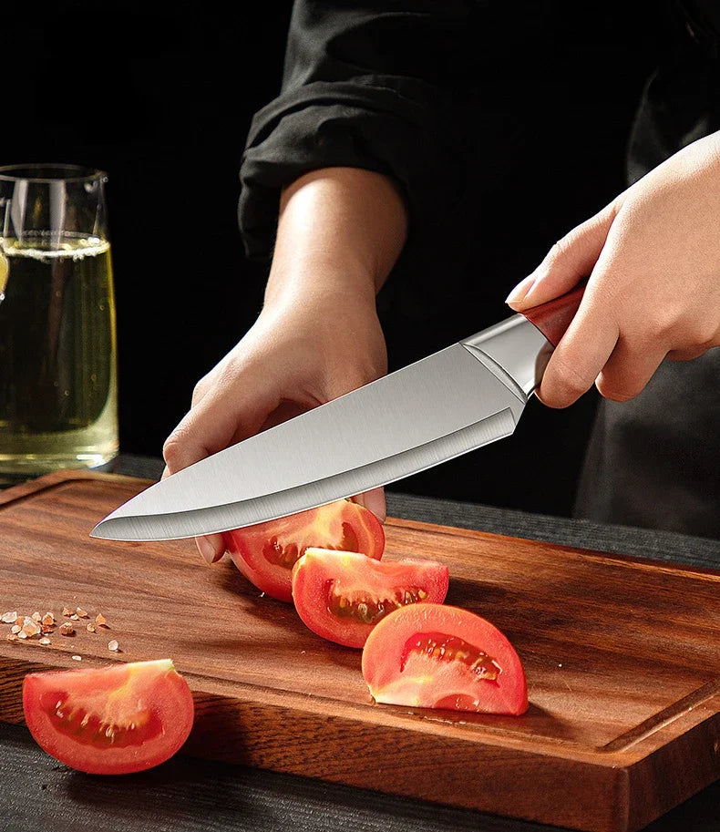 Una mano sosteniendo un cuchillo de despiece de acero inoxidable cortando un tomate en una tabla de cortar de madera.