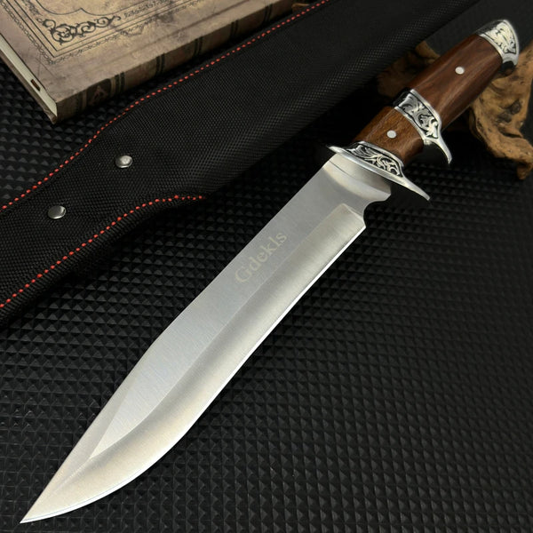 Silberne Camping-Überlebensmesser mit Holzgriff und schwarzem Sheath auf dunkler Oberfläche neben einem alten Buch.